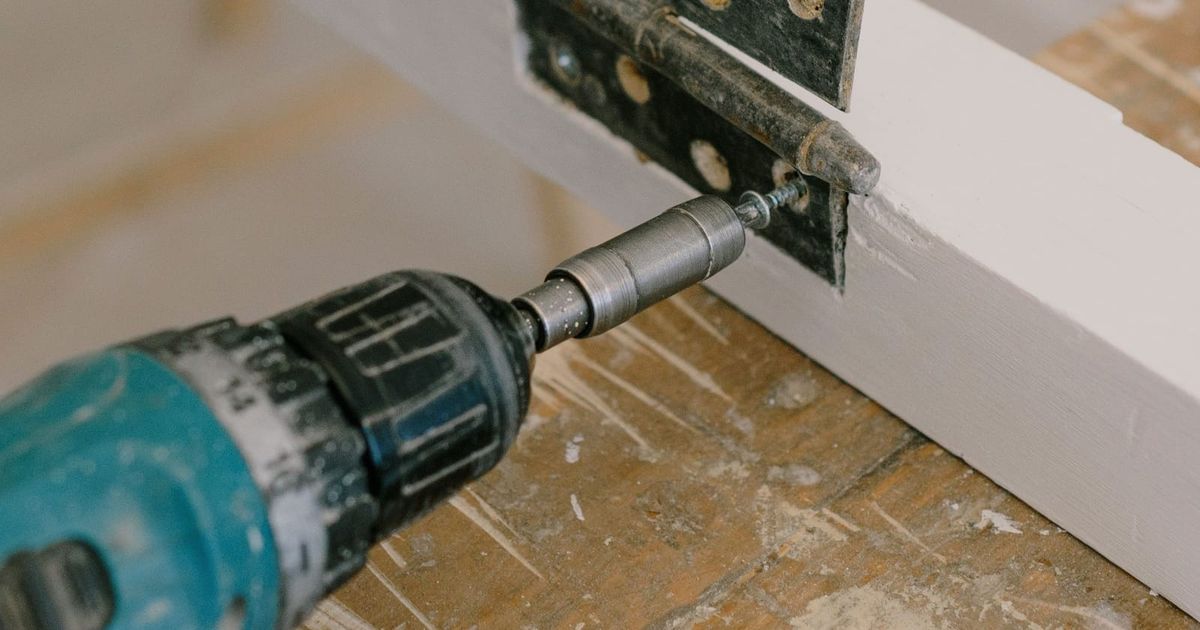 Cordless drill driving a screw into a hinge on a timber frame