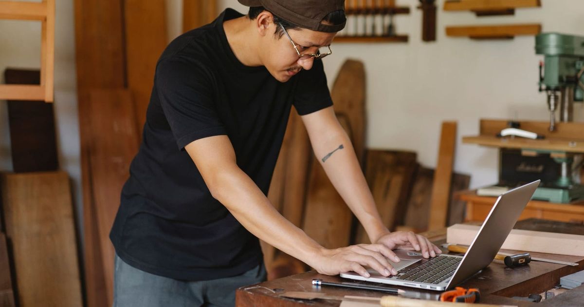 Tradie in a workshop typing on a laptop at the workbench